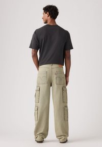 Μαύρο t-shirt με στρογγυλή λαιμόκοψη και ανοιχτοπράσινο παντελόνι cargo με πολλαπλές τσέπες και ξεθωριασμένο τελείωμα, που φαίνεται από πίσω.