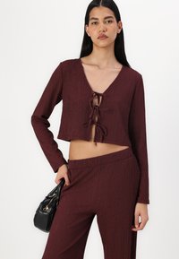 Maroon cropped top with a V-neck, long sleeves, and front tie detail. Textured ribbed fabric. Paired with matching pants and a black handbag.