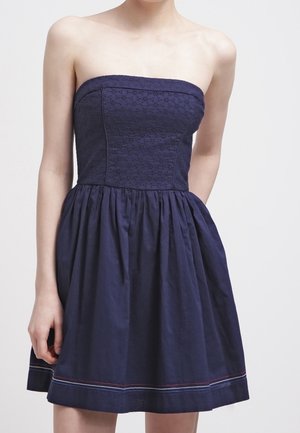 Abito blu navy senza spalline con corpetto ricamato aderente e gonna arricciata che termina sopra le ginocchia, indossato da una persona in piedi.