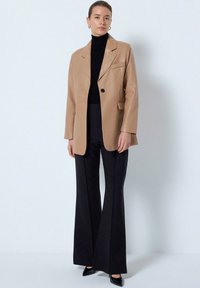 Beige læderblazer med enkelt sort knap, parret med en sort turtleneck og højtaljede, flagrende sorte bukser. Sorte spidse sko.