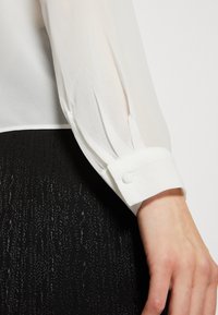 Blusa bianca trasparente con polsini abbottonati, caratterizzata da leggere pieghe. Indossata sopra un tessuto nero texturizzato con motivi verticali.
