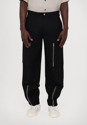 Svart cargo bukser med glidelåsdetaljer på framlommene og mansjettene, med en avslappet passform og avsmalnende ben. Stoffet har en glatt tekstur.