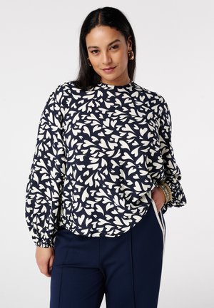 Vrouw met een marineblauwe blouse met witte abstracte hartpatronen, gecombineerd met marineblauwe broek, gouden oorringen en armbanden, poserend met hand in de zak.