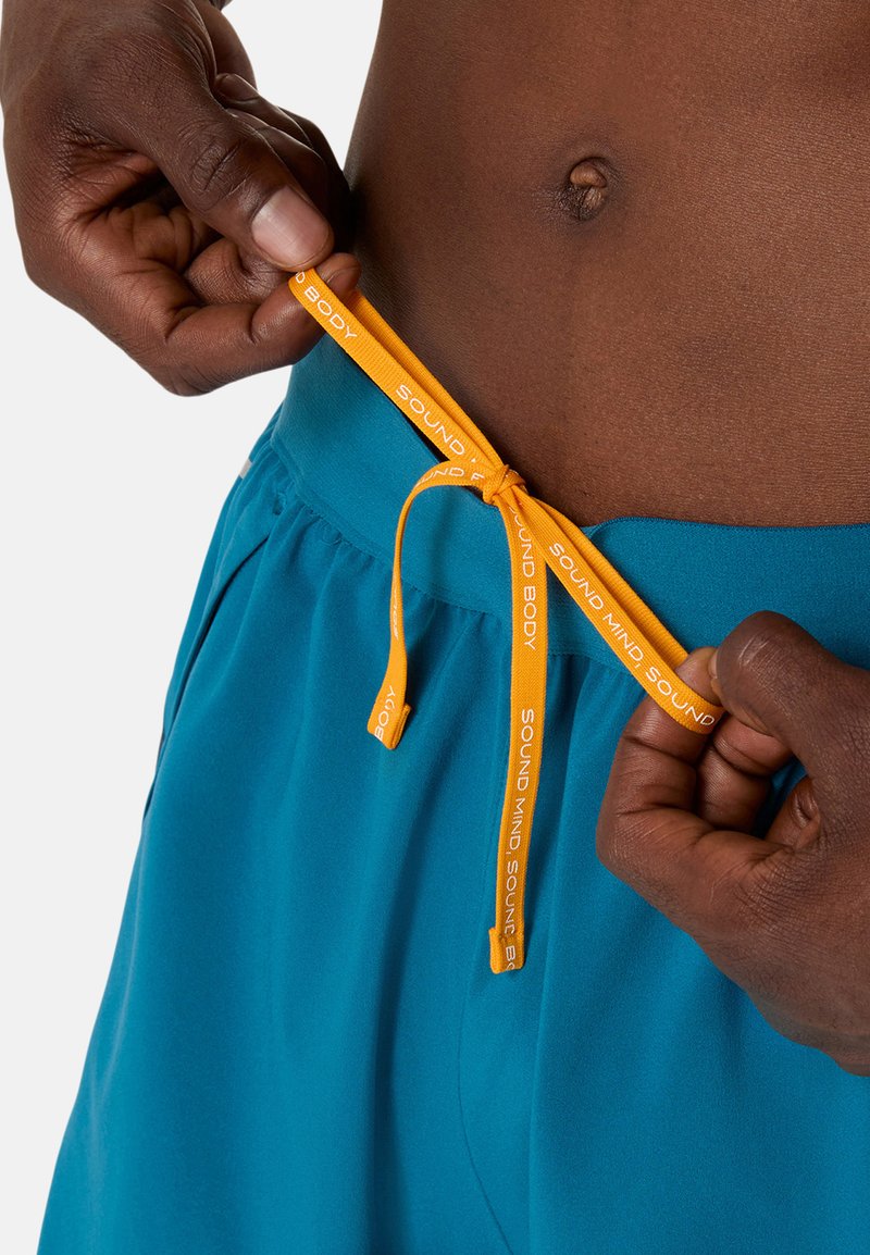 Shorts turquoise avec une taille élastique, dotés d'un cordon de serrage orange portant l'inscription "SOUND MIND SOUND BODY". Tissu lisse, coupe décontractée.