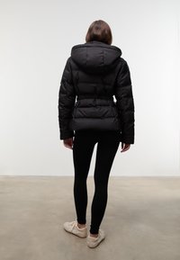 Czarna puchowa kurtka z kapturem, z paskiem w talii i pikowaną fakturą, noszona z czarnymi legginsami i jasnym obuwiem.