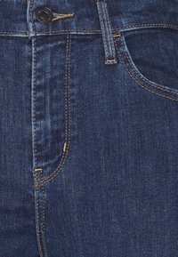 Jeans de denim azul escuro com costura em contraste, bolsos frontais e um fecho de botão visível na cintura. Textura suave com ligeiro brilho.