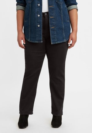 Person iført sorte bukser, sorte ankelstøvler, hvid skjorte og blå denimjakke med op rullede ærmer, stående mod en ensfarvet baggrund.