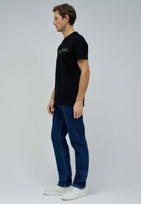 Camiseta de algodón negra con la palabra "JEANS" impresa en gris oscuro, combinada con jeans de mezclilla azules y zapatillas blancas. Vista lateral del modelo contra un fondo gris.