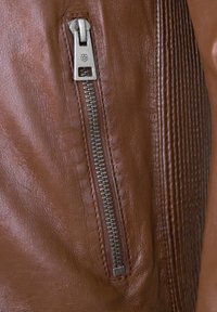 Braune Lederjacke mit silbernem Reißverschluss, strukturiertem Finish und gerippten Details an der Seite. Nahaufnahme des Reißverschlussbereichs.