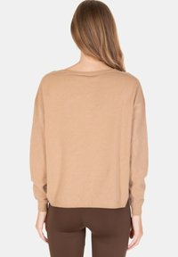 Donna con capelli lunghi che indossa un maglione beige ampio e pantaloni marroni, in piedi di spalle contro uno sfondo chiaro.