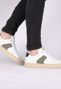 Zapatillas de piel blanca con detalles en oliva, suelas de goma color tostado y cordones redondos, exhibidas en pies que llevan unos jeans ajustados negros.