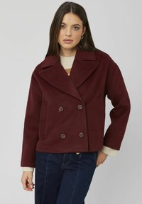 Veste en mélange de laine bordeaux à double boutonnage avec de grands revers, deux poches latérales et boutons marron, associée à un pull crème.