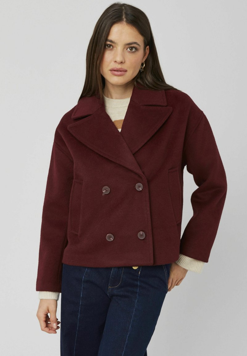 Veste en mélange de laine bordeaux à double boutonnage avec de grands revers, deux poches latérales et boutons marron, associée à un pull crème.