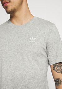 Pelēks īsām piedurknēm T-krekls, izgatavots no mīkstas kokvilnas, ar mazu baltu Adidas logo uz krūškurvja un apaļu kakla izgriezumu.