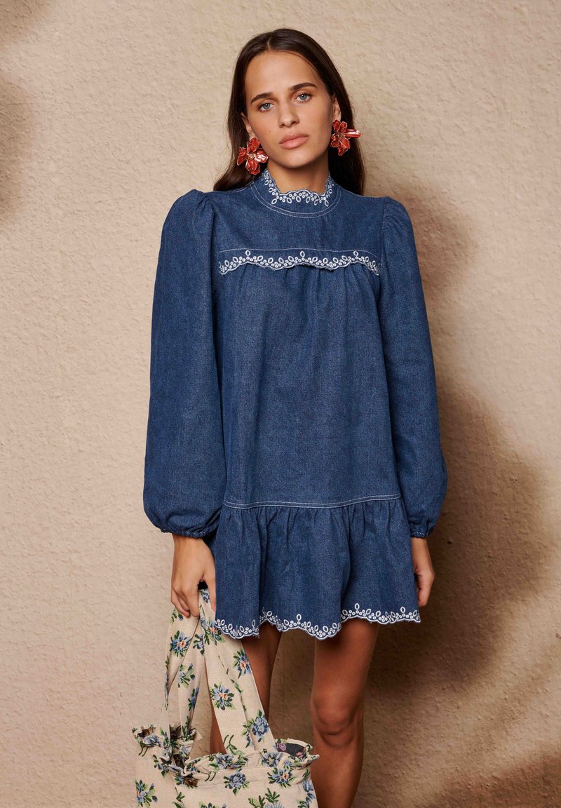 Τζιν φόρεμα με ψηλό γιακά, φουσκωτά μανίκια και κεντημένης σχεδίασης τελείωμα με κυματιστό μοτίβο. Το μοντέλο κρατά μια τσάντα tote με λουλουδάτο μοτίβο. Μονόχρωμο μπλε.