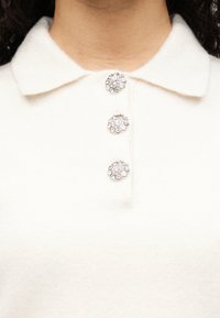 Λευκό πόλο πουκάμισο με απαλή υφή, που διαθέτει γιακά και τρία διακοσμητικά ασημένια κουμπιά με κυκλικά, κρυσταλλώδη σχέδια.