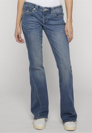 Widok z przodu osoby noszącej niebieskie dżinsy bootcut o średnim stanie z pięcioma kieszeniami i białe trampki, stojącej na tle jasnoszarym.