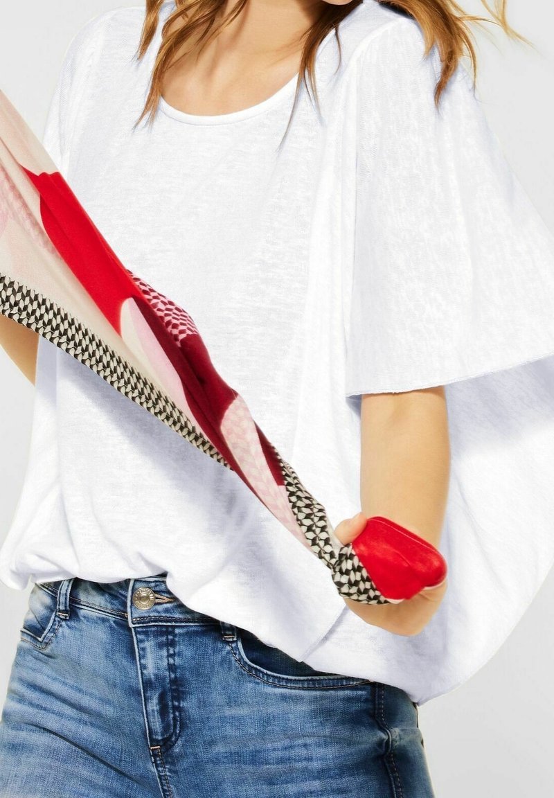 Vit, löst sittande topp med vida ärmar. Bärs med denimjeans. En flerfärgad scarf med mönster i röd och rosa hålls.