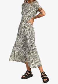 Sukienka midi w kwiatowy nadruk w kolorze czarnym, białym i żółtym. Posiada okrągły dekolt, krótkie rękawy i boczne rozcięcia, luźny krój. Czarne sandały na platformie.