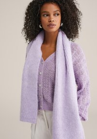 Lila kardigāns ar adītu rakstu, pogām un saskaņotu ribotu šalli. Tekstūrēta auduma, garām piedurknēm un atvērta kakla izgriezums.