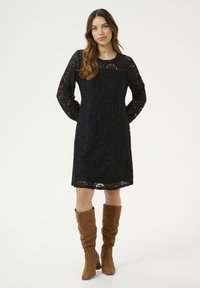 Schwarzes Spitzenkleid mit langen Ärmeln, das mit detaillierten Blumenmustern und einem gewellten Ausschnitt versehen ist. Getragen mit kniehohen braunen Wildlederstiefeln.