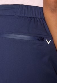 Detailní záběr na tmavě modrou oděv s kapsou na zip a bílým logem, přičemž nad ním je viditelná texturovaná růžová látka.