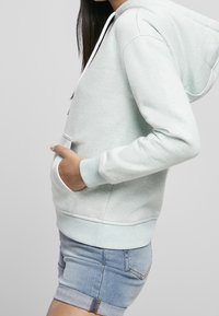 Felpa verde chiaro in tessuto morbido e testurizzato, con tasca a marsupio, cappuccio con cordino e polsini a coste, abbinata a pantaloni corti in denim.