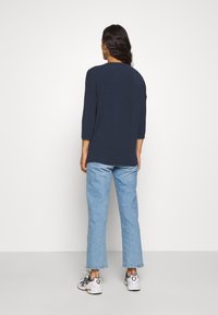 Marineblå topp med 3/4-ermer og avslappet passform. Kombinert med lyseblå rette jeans og hvite joggesko med svarte og oransje detaljer.
