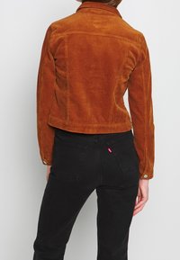 Vrouw in een kort roestkleurig corduroyjack en hoge zwarte spijkerbroek, staand met haar rug naar de camera tegen een eenvoudige achtergrond.