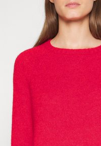 Rode, gebreide trui met een ronde halslijn, lange mouwen en een zachte textuur met een ribbelrand. De close-up benadrukt de kleur en de stofdetails.