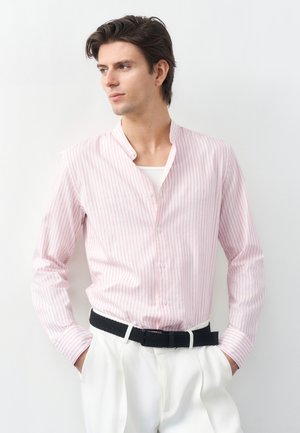 Άνδρας με ροζ και λευκή ριγέ πουκάμισο, μαύρη ζώνη και λευκό παντελόνι, όρθιος με τα χέρια στις τσέπες, κοιτάζει προς το πλάι.