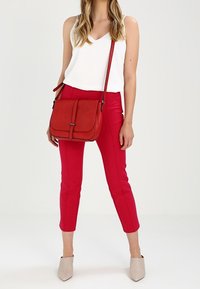 Červená kabelka přes rameno s klopou, nošená přes bílý top bez rukávů a červené přiléhavé kalhoty, doplněná béžovými lodičkami s špičatou špičkou.