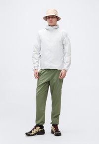 Άντρας που στέκεται φορώντας μπεζ καπέλο τύπου bucket, ανοιχτογκρι μπουφάν με φερμουάρ, χακί παντελόνι και πεζοπορικά παπούτσια σε μπεζ και μαύρο.