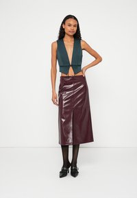 Grünes, kurz geschnittenes Top mit tiefem V-Ausschnitt und Bindeband, kombiniert mit einem glänzenden burgunderroten Midirock mit Frontschlitz. Schwarze Schuhe.