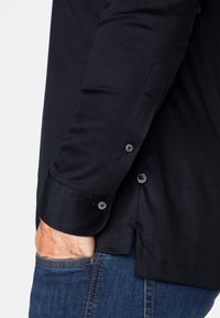 Schwarzes Langarmhemd mit glatter Textur, geknöpften Manschetten und einem seitlichen Schlitz. Kombiniert mit blauer Jeans, die eine lässige Passform zeigt.