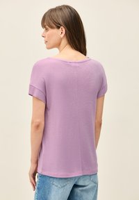 Světlý purpurový top s krátkým rukávem a volným střihem, jemnou strukturou a širokým výstřihem. Zadní část má detail švu a ohrnovací manžety.