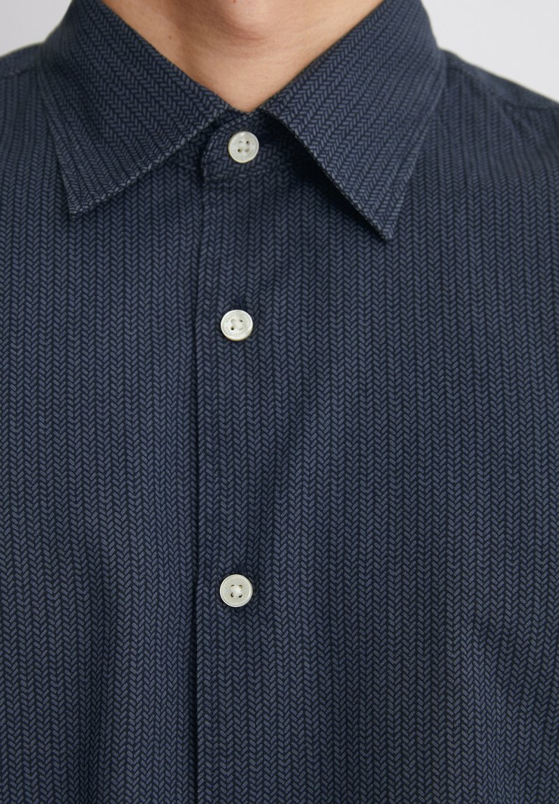 Chemise bleu marine à motifs avec un design en chevron, col pointu et boutons blancs. Texture lisse avec un léger éclat.