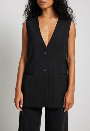 Femme portant un blazer noir sans manches avec trois boutons et deux poches fendues à l'avant, associé à un pantalon noir à jambes larges.