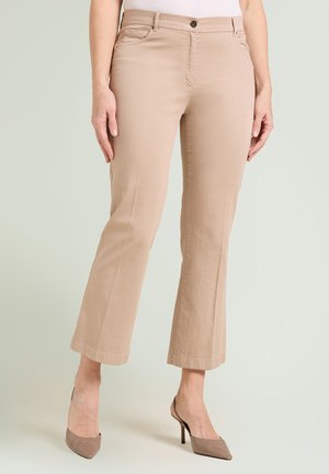 Frau trägt beige verkürzte Hose mit Seitentaschen und taupefarbenen spitzen High Heels, steht vor neutralem Hintergrund.