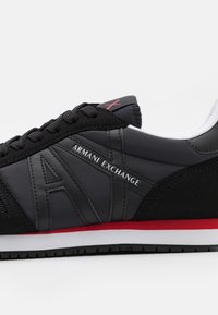 Chaussure de sport noire avec une tige synthétique lisse, des accents en daim, un logo "ARMANI EXCHANGE" bien visible, et des détails rouges sur la semelle.