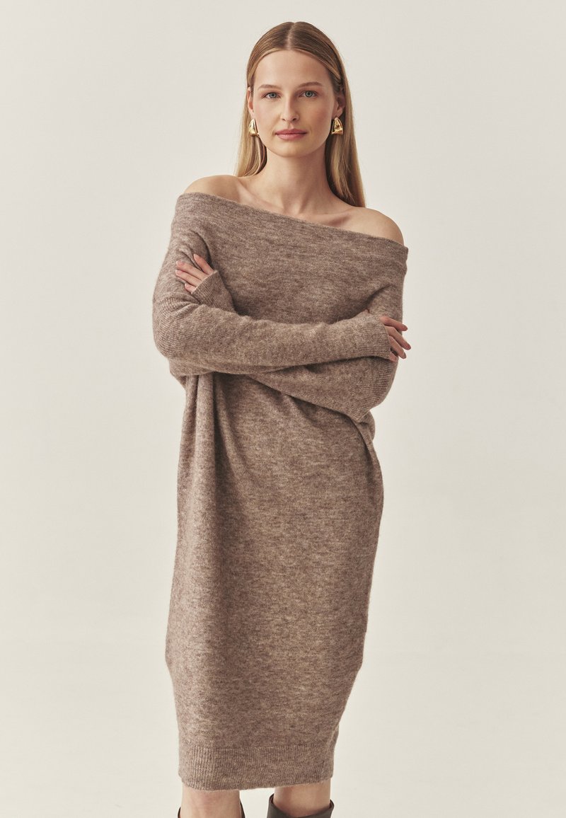 Strickkleid mit schulterfreiem Design aus weichem, leichtem braunem Wollmix, langen Ärmeln und gerippten Bündchen; das Modell steht mit verschränkten Armen.
