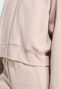 Felpa cropped rosa con chiusura a zip, presenta righe bianche sulle maniche, una texture morbida e un piccolo logo riflettente sul bordo.