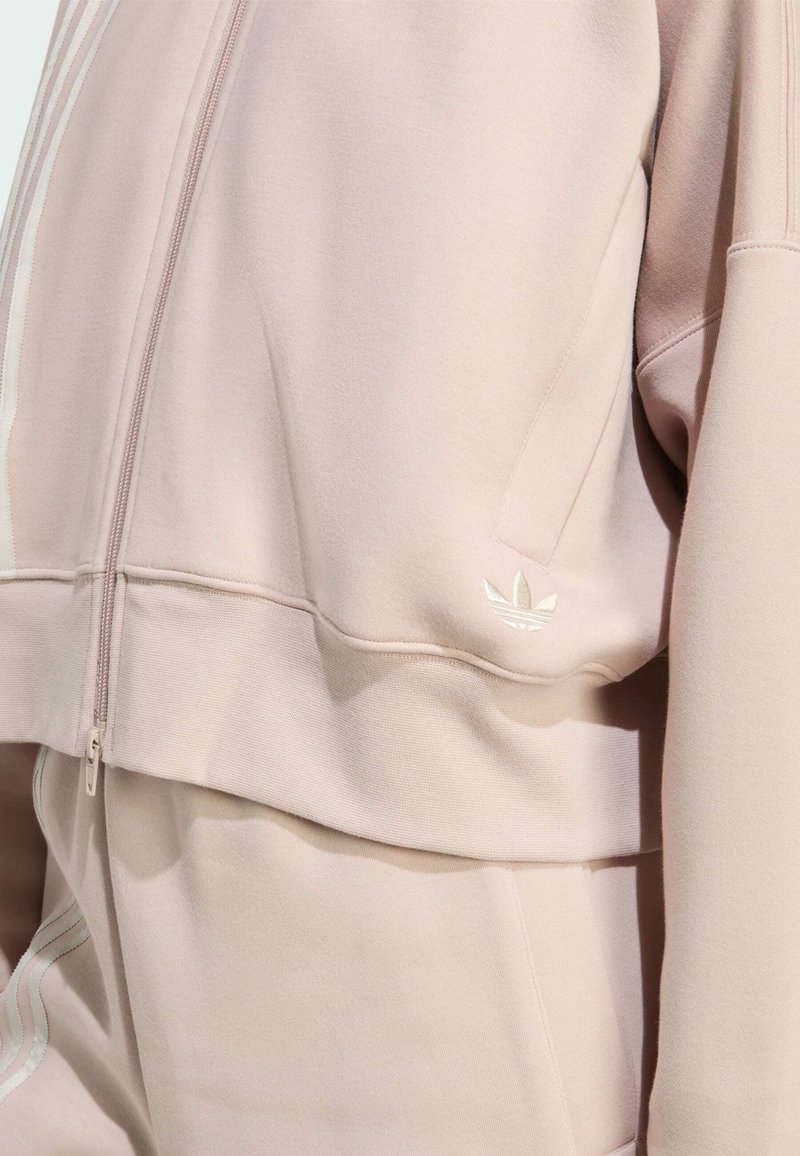 Lyserød cropped sweatshirt med lynlås, har hvide striber på ærmerne, blød tekstur og en lille reflekslogo detalje på kanten.