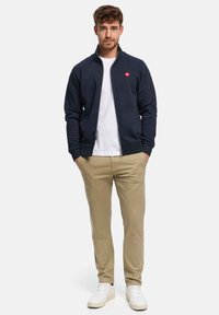 Sweat-shirt à fermeture éclair marine avec col montant et logo rouge, associé à un pantalon beige et des baskets blanches. Texture lisse et coupe décontractée.