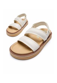 Sandalias de cuero blanco con dos correas anchas, plantilla de color marrón, y una suela de color beige claro con un acento cosido. Diseño plano, con la parte trasera abierta.