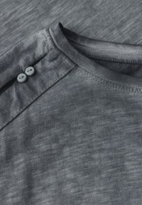Camisa gris con una textura suave, que presenta un cuello acanalado y dos botones gris claro cerca del hombro. Diseño simple, sin patrones.