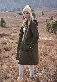 Parka verde oliva con cappuccio, due tasche frontali e chiusura con zip, abbinata a un berretto a maglia color crema con pom-pom in cima.