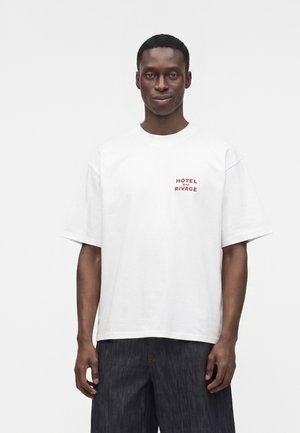 Homme portant un t-shirt blanc oversize avec un texte rouge "Hôtel du Rivage" sur la poitrine et un pantalon large foncé, se tenant devant un fond blanc uni.