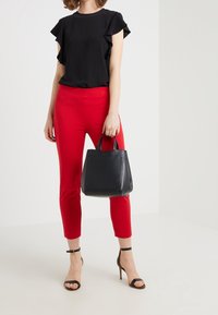 Kvinna som står i en svart topp med volanger, röda åtsittande byxor, svarta ankelsnörade klackar, och håller en svart läderhandväska vid sin sida.