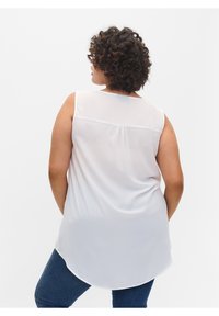 Blusa sin mangas blanca, confeccionada en una tela ligera y transparente, con un dobladillo curvado y detalles de pliegues en la parte posterior, combinada con jeans azules.